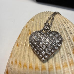 Puede incluir: Un collar con colgante de coraz&oacute;n plateado y cadena. El coraz&oacute;n est&aacute; cubierto de peque&ntilde;as piedras claras y brillantes dispuestas en forma de rejilla. El collar descansa sobre una concha marina beige y marr&oacute;n.