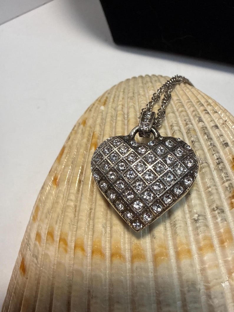 Puede incluir: Un collar con colgante de coraz&oacute;n plateado y cadena. El coraz&oacute;n est&aacute; cubierto de peque&ntilde;as piedras claras y brillantes dispuestas en forma de rejilla. El collar descansa sobre una concha marina beige y marr&oacute;n.