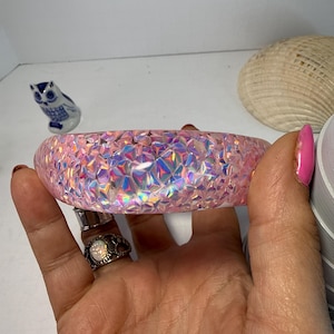 May include: A pink, iridescent bracelet with a geometric pattern. The bracelet is held in a hand, showcasing its sparkling, textured surface. The bracelet is a wide, rounded shape, and the light catches the facets, creating a dazzling effect.
