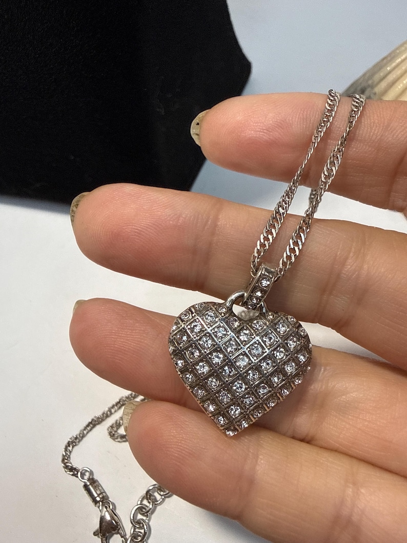 Puede incluir: Un collar con colgante de coraz&oacute;n plateado. El colgante en forma de coraz&oacute;n est&aacute; cubierto de peque&ntilde;as piedras claras y brillantes dispuestas en un patr&oacute;n de rejilla. La cadena del collar es de estilo eslabones finos, con un cierre a juego.