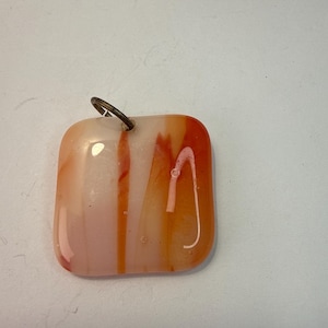 May include: A square pendant with rounded corners, featuring a blend of orange, white, and pink hues. The glass pendant has a metal loop at the top for hanging. The colors are swirled and streaked, creating a unique design.