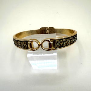 May include: A gold-tone hinged bangle bracelet featuring a black enamel inlay with a repeating gold star and geometric pattern. The bracelet has a polished finish and a central clasp mechanism with two circular loops at the opening.