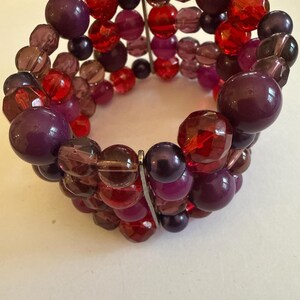 Puede incluir: Una pulsera de cuentas con una mezcla de cuentas moradas y rojas. La pulsera está hecha de cuentas de varios tamaños y formas, incluyendo diseños redondos y facetados. La paleta de colores incluye tonos de morado oscuro, burdeos y rojo rubí.