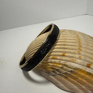 May include: A gold-toned metal bangle bracelet with a textured black band. The bracelet is resting on a large, light brown seashell. The bracelet's design features a sleek, modern aesthetic, suitable for everyday wear or special occasions.