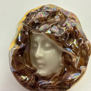 May include: A ceramic brooch featuring a woman's face with flowing hair. The face is a pale color, surrounded by brown hair with iridescent highlights. The brooch has a gold-colored border.