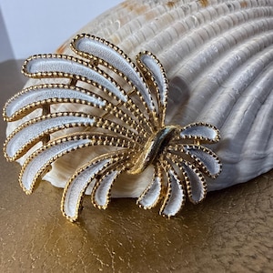 Puede incluir: Broche vintage en tono dorado con diseño de esmalte blanco. La broche tiene forma de abanico con elementos curvos y en capas. Los bordes están detallados con pequeñas cuentas doradas. La broche está sobre una concha marina.