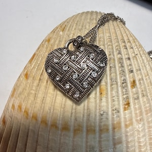 Puede incluir: Collar con colgante de coraz&oacute;n plateado con un dise&ntilde;o texturizado y peque&ntilde;as gemas transparentes. El colgante en forma de coraz&oacute;n cuelga de una delicada cadena. El collar se muestra sobre una gran concha texturizada.