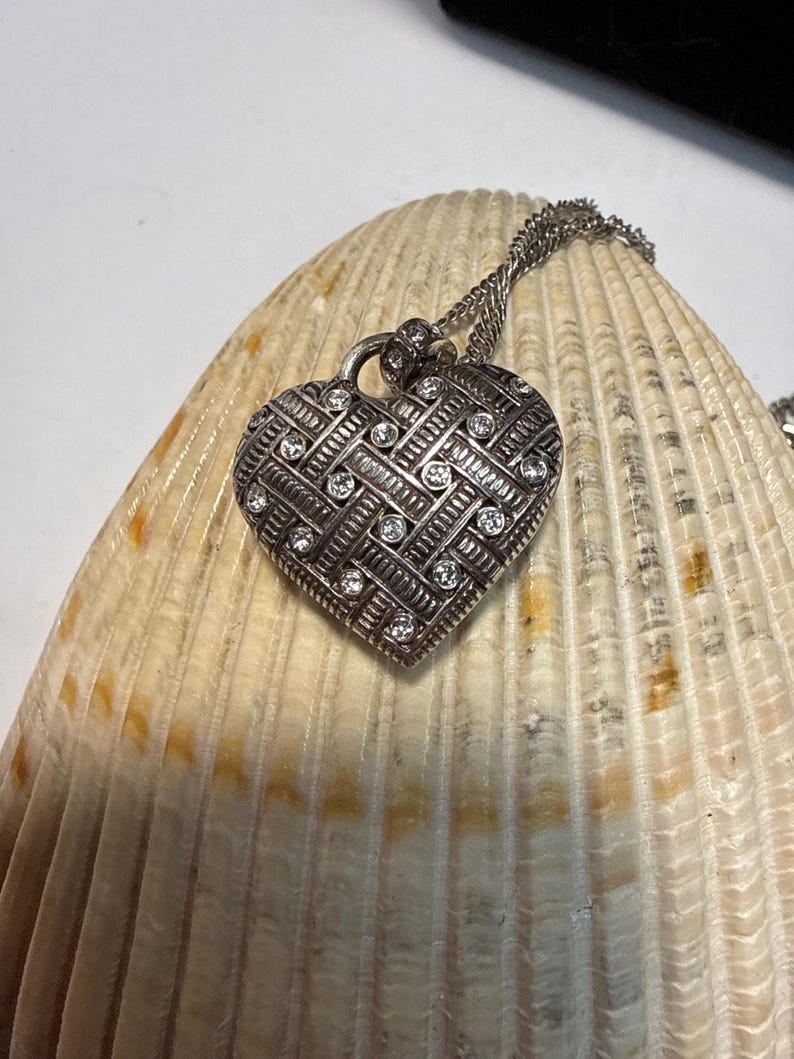 Puede incluir: Collar con colgante de coraz&oacute;n plateado con un dise&ntilde;o texturizado y peque&ntilde;as gemas transparentes. El colgante en forma de coraz&oacute;n cuelga de una delicada cadena. El collar se muestra sobre una gran concha texturizada.