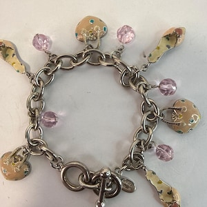 Pode incluir: Pulseira de berloques prateada com fecho de alternância e vários berloques. Os berloques incluem chinelos rosa e amarelos, berloques em forma de cogumelo e detalhes de contas rosa. Um acessório caprichoso.