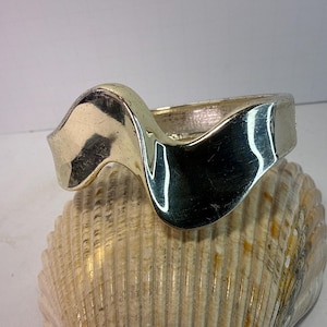 Può includere: Un braccialetto rigido in metallo argentato con un design a onda. Il braccialetto è appoggiato su una conchiglia beige. Il braccialetto ha una superficie lucida e riflettente.