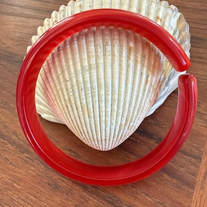 May include: A vibrant red bangle bracelet with a smooth, glossy finish. The open-ended bracelet is displayed on a large, textured seashell, set against a warm, wooden surface. The bracelet's bold color and simple design make it a striking accessory.