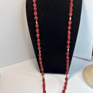 May include: A long necklace featuring red, twisted beads interspersed with small, gold-colored beads. The necklace is displayed on a black mannequin, showcasing its length and design. The beads have a glossy finish.