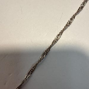 Puede incluir: Una cadena plateada con un dise&ntilde;o de cuerda trenzada. La cadena se muestra en diagonal sobre un fondo claro. Los eslabones est&aacute;n tejidos apretadamente, creando una apariencia texturizada. Esta joya es adecuada para diversas ocasiones.