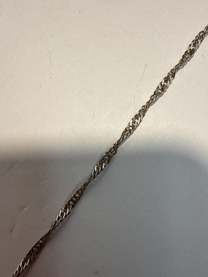 Puede incluir: Una cadena plateada con un dise&ntilde;o de cuerda trenzada. La cadena se muestra en diagonal sobre un fondo claro. Los eslabones est&aacute;n tejidos apretadamente, creando una apariencia texturizada. Esta joya es adecuada para diversas ocasiones.