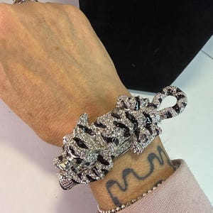 May include: A silver-toned bracelet in the shape of a tiger, encrusted with clear crystals and black enamel stripes. The bracelet is worn on a wrist with a tattoo and a silver beaded bracelet. A matching ring is also visible.