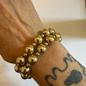 May include: A double-strand bracelet composed of polished gold-tone spherical beads. The design features larger metallic spheres interspersed with smaller beads, creating a layered appearance around the wrist with a reflective, smooth finish.