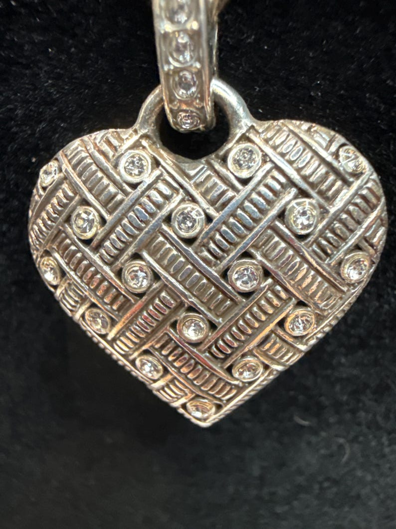 Puede incluir: Colgante plateado en forma de coraz&oacute;n con un dise&ntilde;o tejido y gemas claras incrustadas. El colgante tiene una superficie texturizada y un lazo en la parte superior para una cadena. El coraz&oacute;n es una forma cl&aacute;sica.