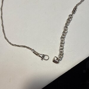 Puede incluir: Un collar plateado con un dise&ntilde;o de cadena de cuerda trenzada. El collar presenta un cierre de mosquet&oacute;n y una cadena ajustable con un dije en forma de coraz&oacute;n. El collar se muestra sobre una superficie blanca.