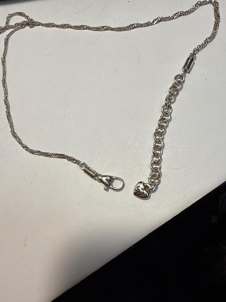 Puede incluir: Un collar plateado con un dise&ntilde;o de cadena de cuerda trenzada. El collar presenta un cierre de mosquet&oacute;n y una cadena ajustable con un dije en forma de coraz&oacute;n. El collar se muestra sobre una superficie blanca.