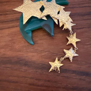 May include: A gold-colored starburst brooch with a textured finish, attached to a dark green, crescent-shaped base. The brooch features a cascade of dangling stars, creating a celestial design. The brooch is likely made of metal.