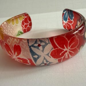 Könnte beinhalten: Ein klarer, offener Armreif mit floralem Muster. Das Armband hat ein rotes, weißes und blaues Blumendesign. Die Blumen sind vor einem roten Hintergrund angeordnet. Das Armband besteht aus einem klaren, glänzenden Material.