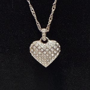Puede incluir: Collar con colgante de coraz&oacute;n plateado. El colgante en forma de coraz&oacute;n est&aacute; incrustado con peque&ntilde;os cristales transparentes dispuestos en un patr&oacute;n de rejilla. La cadena es de dise&ntilde;o trenzado. Fondo negro.