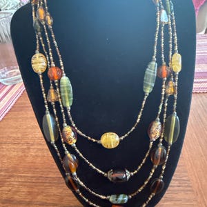 May include: A multi-strand necklace with a variety of beads in shades of brown, amber, green, and yellow. The beads are strung on delicate gold-colored chains, creating a layered effect. The necklace is displayed on a black mannequin.