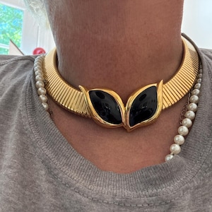 Könnte beinhalten: Eine goldfarbene Metall-Halskette im Choker-Stil mit gerippter Textur und einem zentralen Blickfang aus zwei glatten, schwarzen, mandelförmigen Cabochons in Goldfassungen. Eine separate Kette aus weißen Perlen liegt unter dem Choker.
