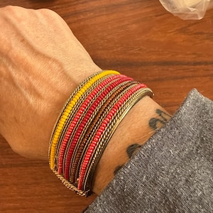 May include: A gold-toned bangle bracelet with rows of colorful beads in shades of yellow, red, and orange. The bracelet is worn on a wrist, with a gray sleeve visible in the frame. The bracelet's design is intricate and eye-catching.