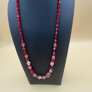 以下が含まれることがあります： ファセットカットされた赤いガラスビーズと、光を反射する虹色のビーズが交互に連なるロングネックレス。ビーズは形や大きさが異なり、ストランドに沿って質感のある外観を作り出しており、黒いトルソーにディスプレイされています。