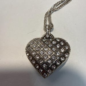 Puede incluir: Collar con colgante de coraz&oacute;n plateado. El colgante en forma de coraz&oacute;n est&aacute; incrustado con numerosas piedras preciosas peque&ntilde;as y transparentes dispuestas en un patr&oacute;n de rejilla. La cadena es de dise&ntilde;o retorcido. Una joya rom&aacute;ntica.