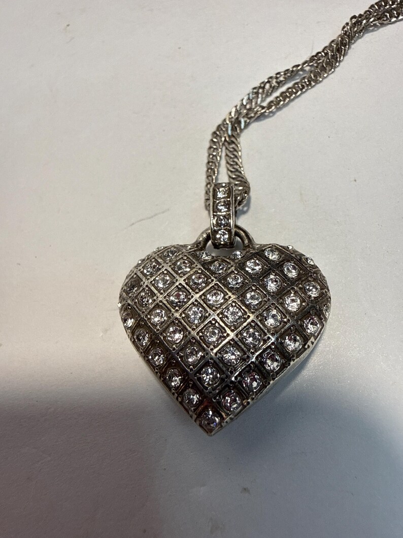 Puede incluir: Collar con colgante de coraz&oacute;n plateado. El colgante en forma de coraz&oacute;n est&aacute; incrustado con numerosas piedras preciosas peque&ntilde;as y transparentes dispuestas en un patr&oacute;n de rejilla. La cadena es de dise&ntilde;o retorcido. Una joya rom&aacute;ntica.