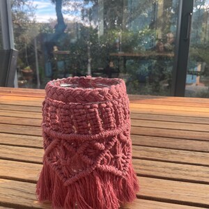 Peut inclure: Un cache-pot en macramé rose poussiéreux avec un motif géométrique et des franges. Le cache-pot est fait de coton épais tissé et est conçu pour s'adapter autour d'un pot en verre. Il est posé sur une table en bois.