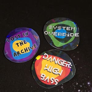 May include: Three holographic stickers with text. One reads "LOST IN THE ARCHIVE" in white on a blue, yellow, and purple background. Another says "SYSTEM OVERRIDE" in white on a green and black background. The third says "DANGER: HIGH BASS" in white on a red, yellow, and orange background.
