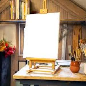 Puede incluir: Un caballete de madera con un lienzo blanco en blanco se encuentra sobre una mesa de madera. Una pequeña maceta de cerámica contiene pinceles, y un tarro de vidrio está lleno de más pinceles. El fondo incluye una estantería con libros y materiales de arte.