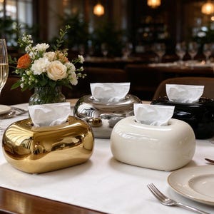 May include: Four oval-shaped tissue box covers in gold, silver, white, and black finishes. Each container features a smooth, rounded design with a central opening for dispensing white tissues, arranged on a table with glasses and flowers.