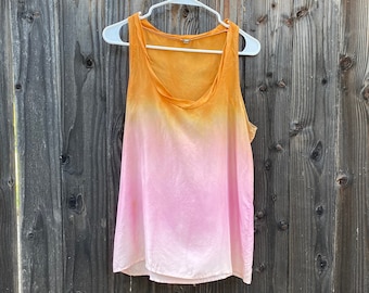 Pode incluir: Top sem mangas com design ombré, que vai do laranja na parte superior ao rosa na inferior. O top está pendurado num cabide branco contra uma cerca de madeira desgastada.
