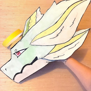 May include: Hand-drawn dragon head mask with a light green base, yellow and black accents, and red eyes. The mask is made of paper and is held by a person's hand. The mask is a unique piece of art.