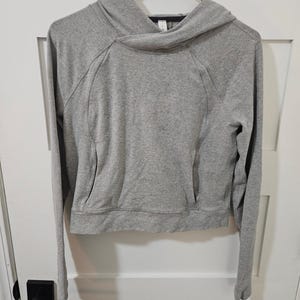 Op de afbeelding: Een grijze hoodie met lange mouwen en een voorzak. De hoodie hangt aan een zwarte hanger tegen een witte deur. De capuchon heeft een uniek, overlappend ontwerp.