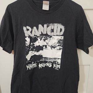 Puede incluir: Camiseta negra con el nombre de la banda "RANCID" en letras blancas en bloque sobre un gráfico en blanco y negro de un coche. Las palabras "WHITE KNUCKLE RIDE" están impresas debajo en blanco.