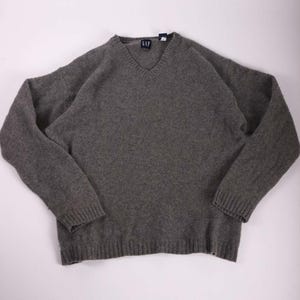 May include: A gray V-neck sweater with long sleeves. The sweater has a ribbed hem and cuffs. The GAP logo is visible near the neckline. The sweater is made of a soft, knitted material.