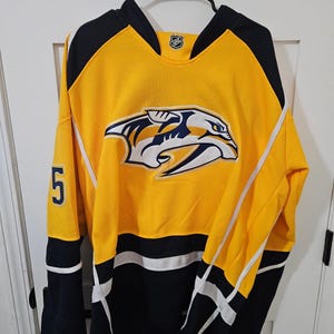 May include: A yellow hockey jersey with black and white accents. The jersey features the Nashville Predators logo on the front and the number 5 on the sleeve. The jersey is hanging on a black hanger.