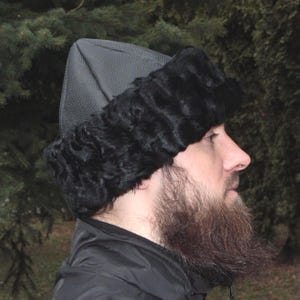 May include: Black fur hat with a leather top. The hat features a textured leather crown and a fluffy fur trim around the ears and forehead. The hat is worn by a person with a beard.