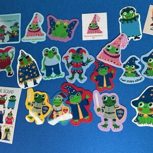 May include: A collection of colourful stickers featuring cartoon frogs in various costumes, including wizards, knights, and jesters. Some stickers include text such as "The horror persists but so do I," "Tiny steps count," "Protect your magic," and "Frog Squad."