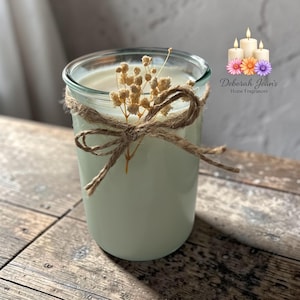 May include: A light green candle in a clear glass jar, adorned with a twine bow and dried flowers. The candle is on a wooden surface. The Deborah Jean's Home Fragrances logo is in the background.