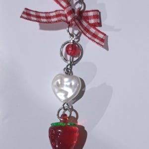 Puede incluir: Un dije decorativo con un lazo de cinta de cuadros vichy rojos y blancos en la parte superior, seguido de un anillo plateado con una pequeña cuenta roja, una cuenta blanca en forma de corazón y un dije de fresa roja translúcida con detalles verdes.