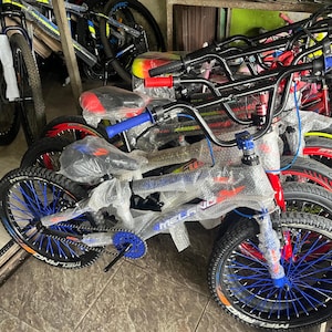 Może przedstawiać: Kolekcja nowych rowerów, w tym rower w stylu BMX z niebieskimi i czarnymi akcentami. Rowery mają czarne opony, czerwone i niebieskie kierownice i są owinięte w ochronny plastik. Koła mają niebieskie szprychy.