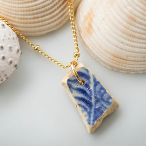 May include: A gold chain necklace with a rectangular pendant made from a piece of blue and white ceramic. The necklace is displayed with two seashells and a white sea urchin shell.