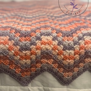 May include: A handmade crochet blanket in a granny square pattern. The blanket features a color gradient of peach, lavender, and cream hues. The blanket has a scalloped edge. The Thistleberry Crochet logo is in the upper right corner.