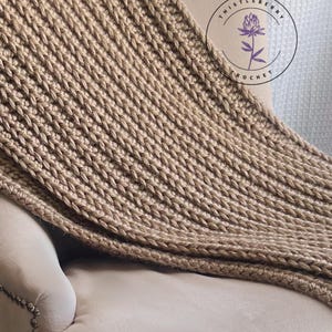 May include: A tan-colored, chunky knit throw blanket draped over the arm of a beige armchair. The blanket has a ribbed texture and is made of a thick yarn. The Thistleberry Crochet logo is visible in the upper right corner.